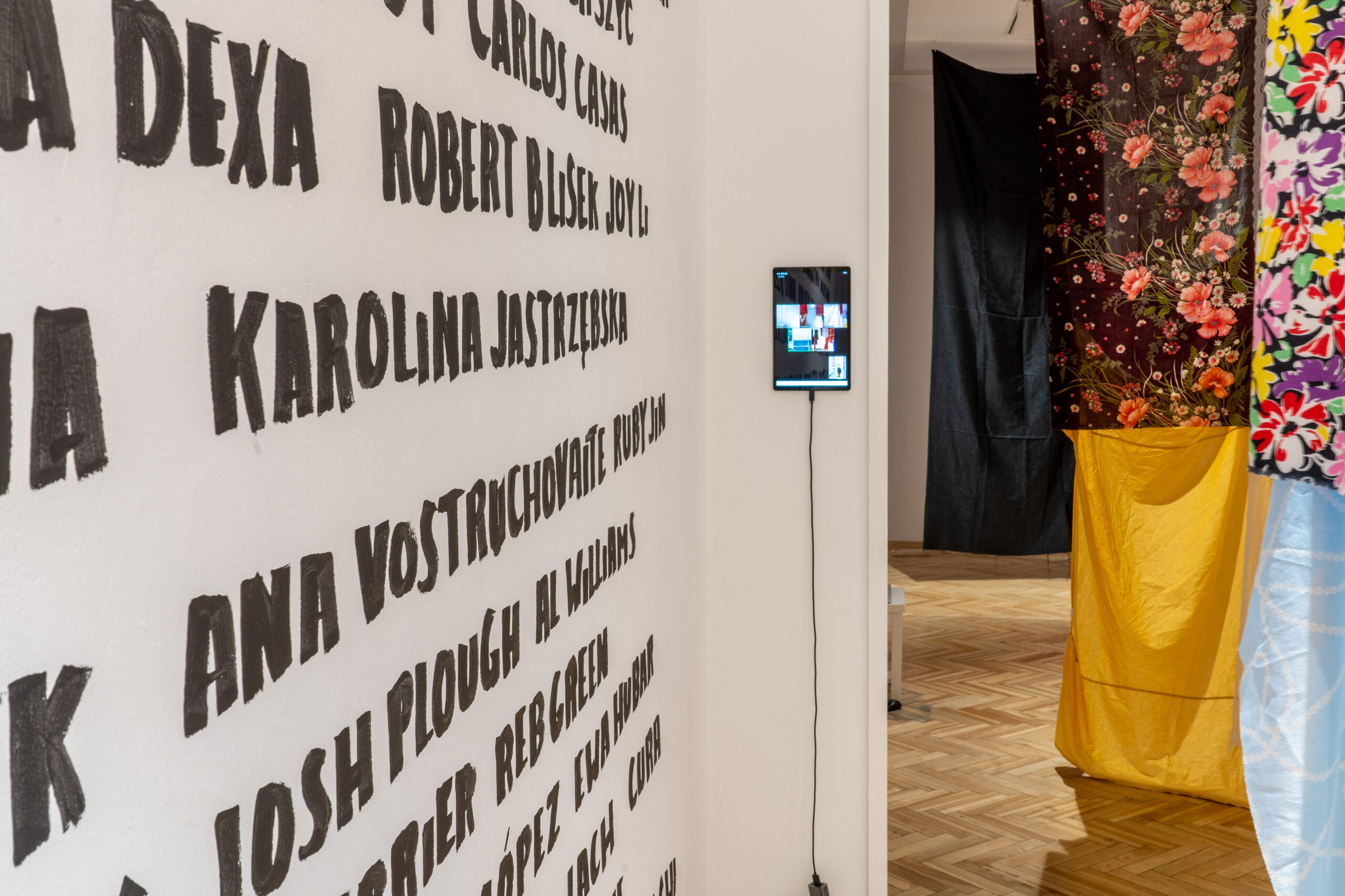 Na zdjęciu znajduje się kawałek ściany z wypisanymi nazwiskami, przyklejony do ściany tablet, wyświetlający konferencję na zoomie oraz fragment wnętrza sali wystawowej z zawieszonymi wewnątrz materiałami.